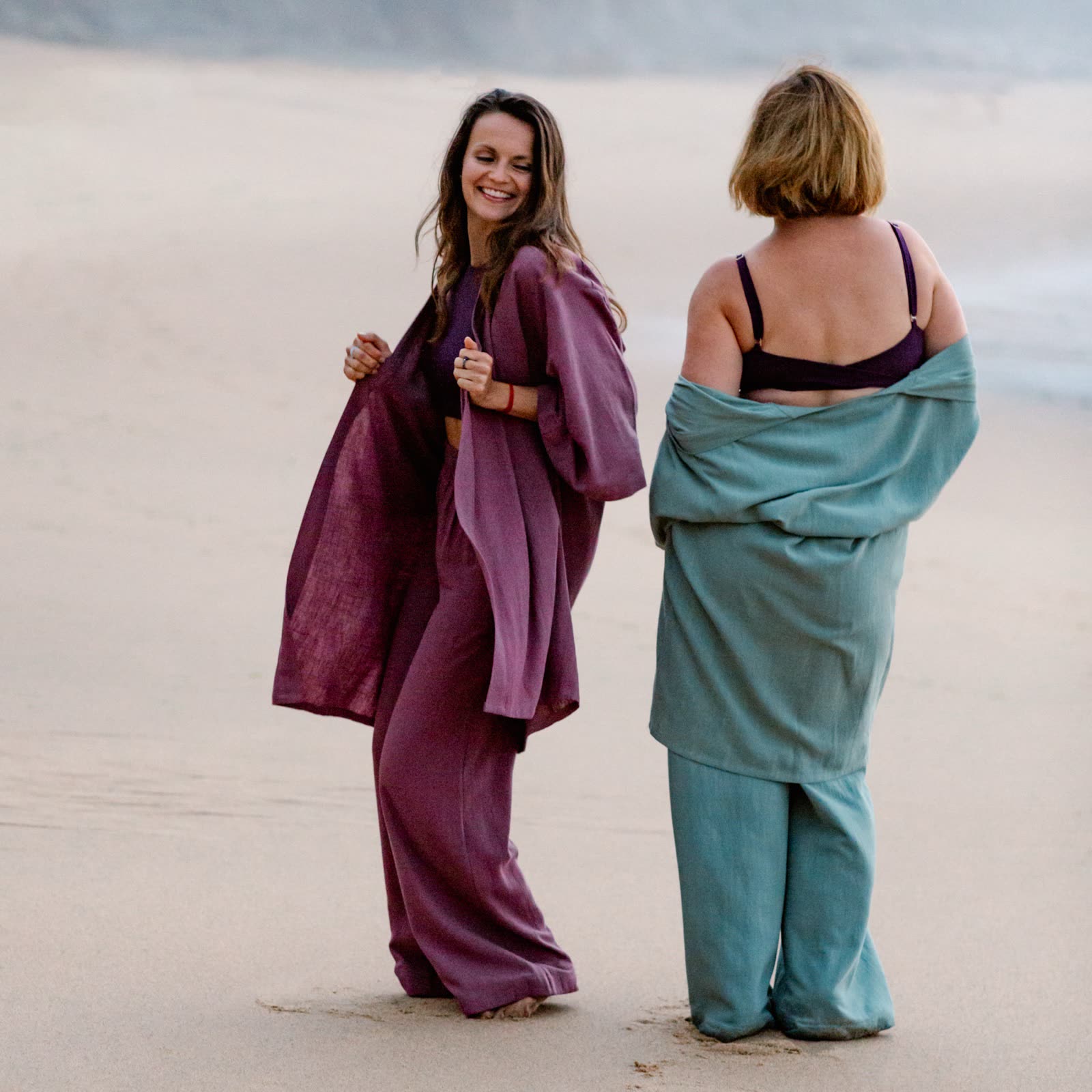 Two women on a beach at dusk, laughing, wrapped in linen
